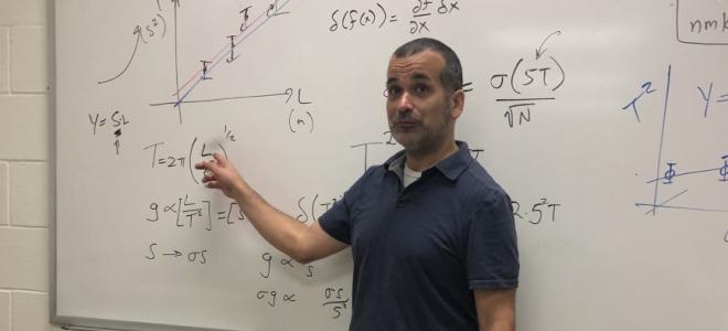 A professor pointing to a white board with equations on it.